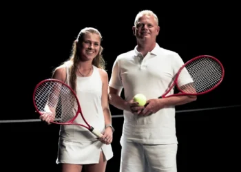 Photo of Courtney Webeck and Mick Leigh dressed in white tennis clothes, holding tennis rackets next to tennis net, with black background