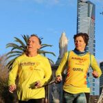 vision impaired runner and their guide are running together using a tether, in Achilles tops.