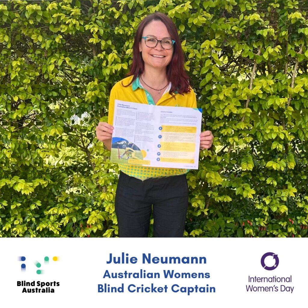 Photo of Julie Neumann wearing green eyeglasses standing outside against a leafy green background holding up a printed double page spread from the Blind Cricket Guide. She is wearing her green and gold Australian IBSA World Games uniform polo, and has long dark hair over her shoulders