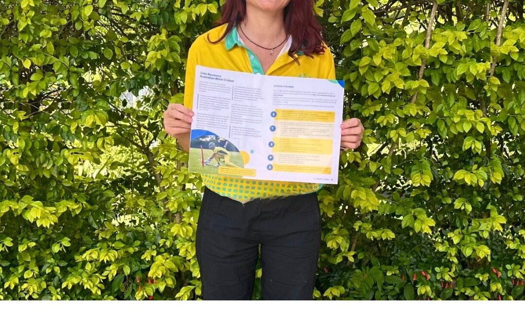 Photo of Julie Neumann wearing green eyeglasses standing outside against a leafy green background holding up a printed double page spread from the Blind Cricket Guide. She is wearing her green and gold Australian IBSA World Games uniform polo, and has long dark hair over her shoulders
