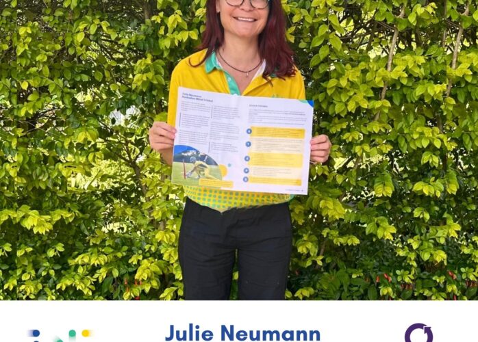 Photo of Julie Neumann wearing green eyeglasses standing outside against a leafy green background holding up a printed double page spread from the Blind Cricket Guide. She is wearing her green and gold Australian IBSA World Games uniform polo, and has long dark hair over her shoulders
