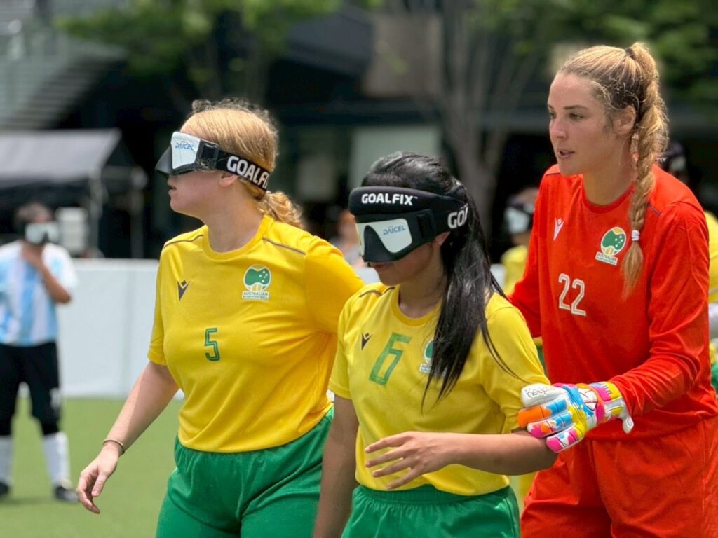 two Australian women's blind football team players and the goal keeper standing on field during a match