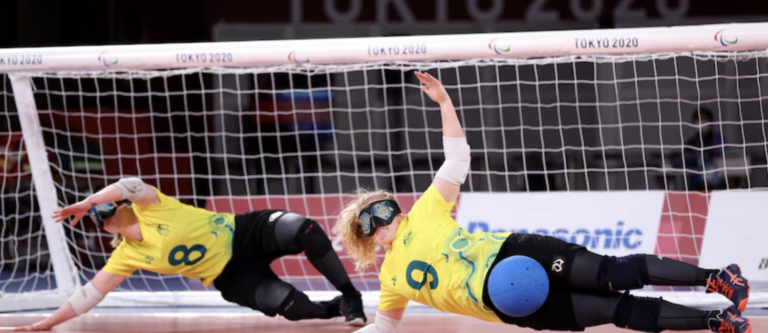 Two Australian goalball players in yellow jerseys dive sideways in front of the goal to block a shot during the Tokyo 2020 Paralympic Games. Both athletes wear eyeshades, knee pads, and elbow pads, with one player (#9) deflecting a blue ball with their body.