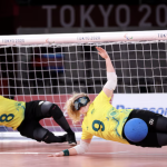 Two Australian goalball players in yellow jerseys dive sideways in front of the goal to block a shot during the Tokyo 2020 Paralympic Games. Both athletes wear eyeshades, knee pads, and elbow pads, with one player (#9) deflecting a blue ball with their body.