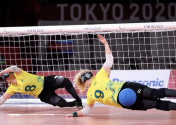 Two Australian goalball players in yellow jerseys dive sideways in front of the goal to block a shot during the Tokyo 2020 Paralympic Games. Both athletes wear eyeshades, knee pads, and elbow pads, with one player (#9) deflecting a blue ball with their body.