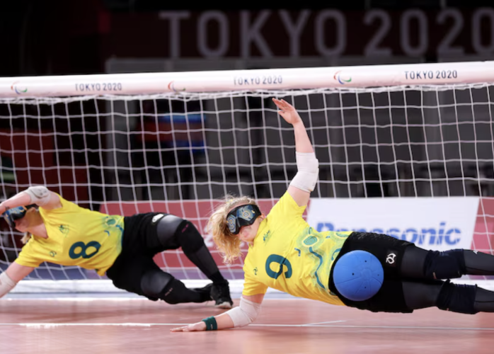 Two Australian goalball players in yellow jerseys dive sideways in front of the goal to block a shot during the Tokyo 2020 Paralympic Games. Both athletes wear eyeshades, knee pads, and elbow pads, with one player (#9) deflecting a blue ball with their body.