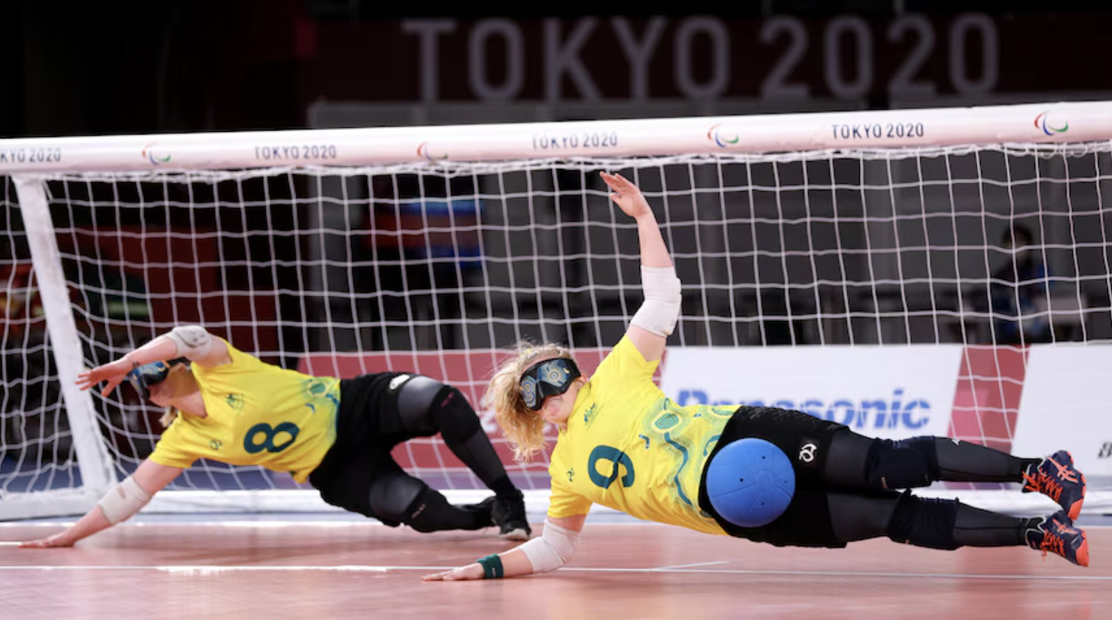 Two Australian goalball players in yellow jerseys dive sideways in front of the goal to block a shot during the Tokyo 2020 Paralympic Games. Both athletes wear eyeshades, knee pads, and elbow pads, with one player (#9) deflecting a blue ball with their body.