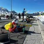 Taryn lies on a shooting mat at a biathlon range, aiming with an adapted rifle while wearing ear protection and a yellow vest. Other athletes train nearby against a backdrop of snow-covered mountains and blue sky.