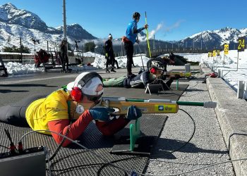 Taryn lies on a shooting mat at a biathlon range, aiming with an adapted rifle while wearing ear protection and a yellow vest. Other athletes train nearby against a backdrop of snow-covered mountains and blue sky.