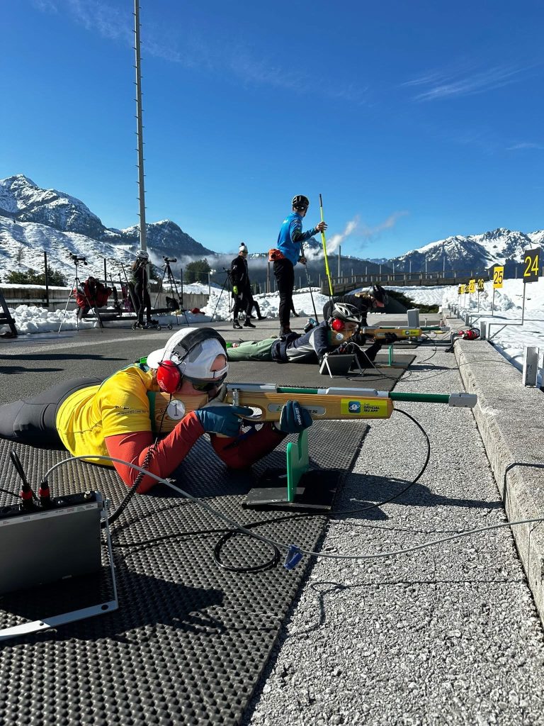 Taryn lies on a shooting mat at a biathlon range, aiming with an adapted rifle while wearing ear protection and a yellow vest. Other athletes train nearby against a backdrop of snow-covered mountains and blue sky.