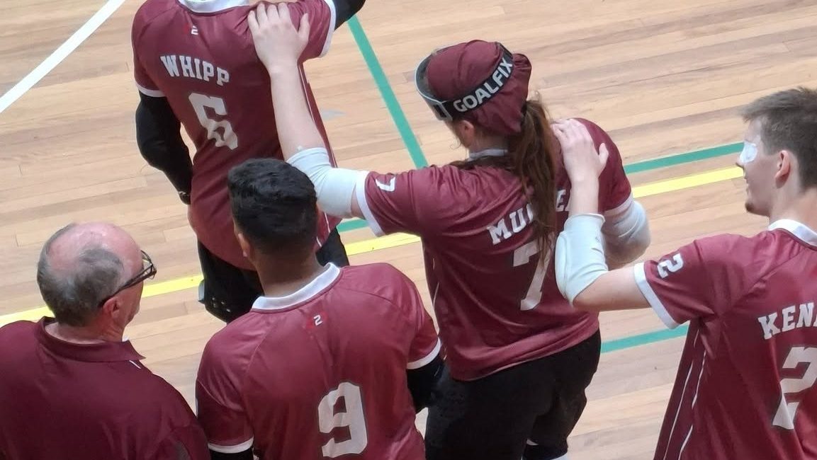 Four goalball players in maroon jerseys stand together at the edge of an indoor court, each with a hand on a teammate’s shoulder. One player wears a cap marked “GOALFIX,” and another has an eye patch visible. A man in a maroon polo stands beside them.