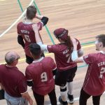 Four goalball players in maroon jerseys stand together at the edge of an indoor court, each with a hand on a teammate’s shoulder. One player wears a cap marked “GOALFIX,” and another has an eye patch visible. A man in a maroon polo stands beside them.