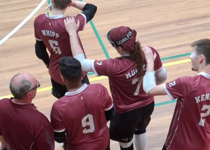 Four goalball players in maroon jerseys stand together at the edge of an indoor court, each with a hand on a teammate’s shoulder. One player wears a cap marked “GOALFIX,” and another has an eye patch visible. A man in a maroon polo stands beside them.